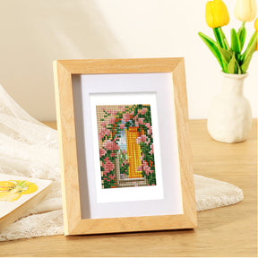 Framed mini diamond painting of floral design on table next to vase of yellow tulips.