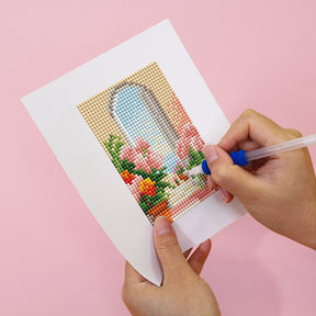 Person crafting a mini diamond painting of a floral window scene on a pink background