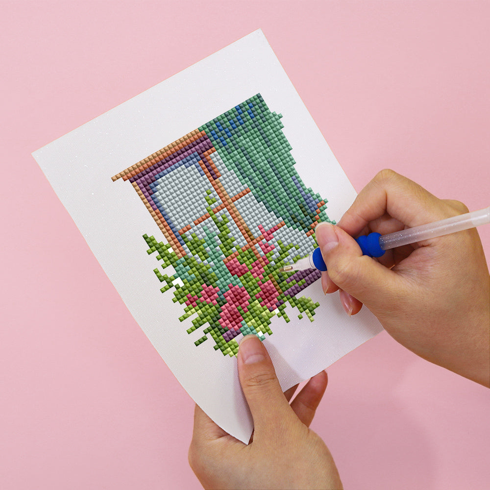 Hands creating FloraleFenster mini art with diamond painting, showcasing vibrant pink flowers and greenery near a window.