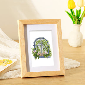 Framed FloraleFenster diamond painting set on a wooden table with flowers and lace fabric in the background.