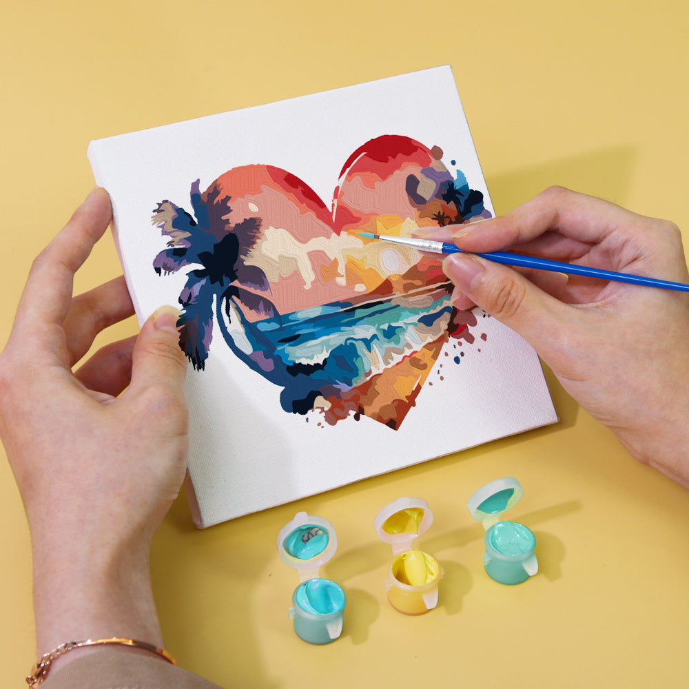 Person painting a tropical heart-shaped scene on a canvas with paint pots on a yellow surface - Mini Kunst Malen nach Zahlen